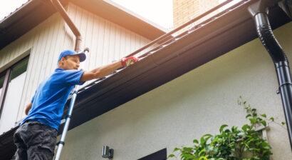 man on ladder cleaning home gutters filled with leaves