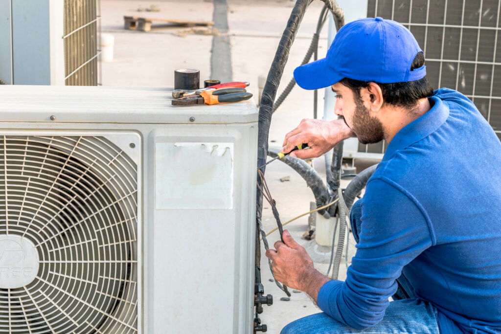 An HVAC technician with a screwdriver conducting a repair on the outdoor unit of an HVAC system.