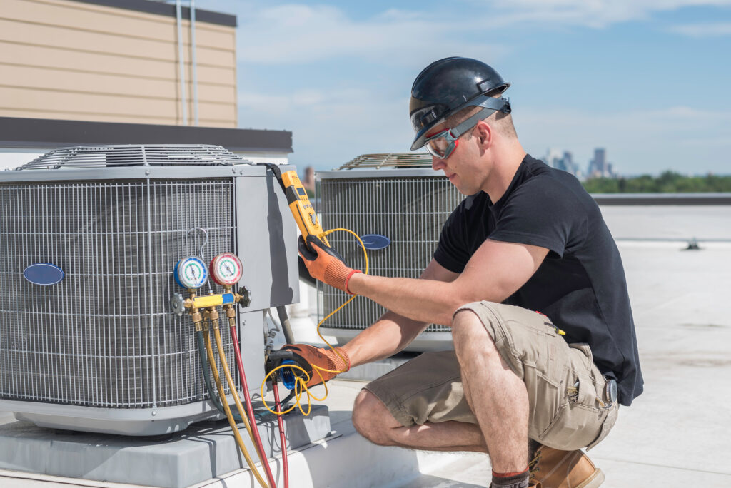 An HVAC professional with a hard hat on kneeling down to check the pressure of an HVAC unit