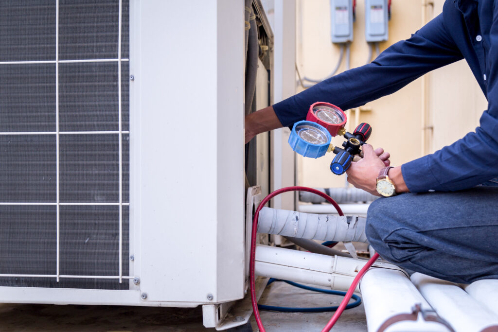 An HVAC technician kneeling down next to a large outdoor HVAC unit checking the pressure.