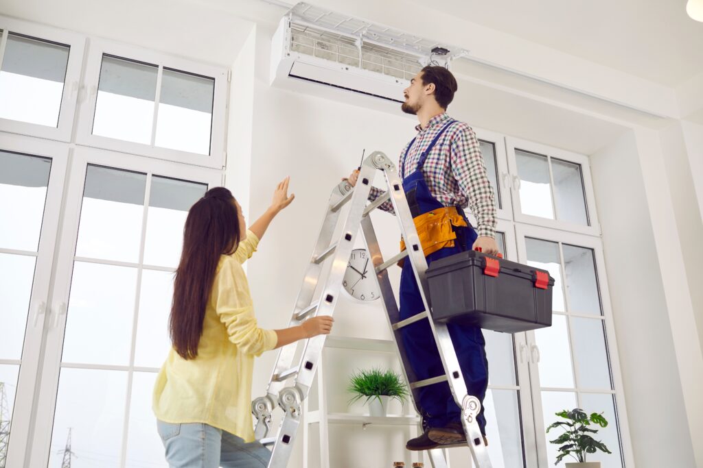 An HVAC technician on a ladder checking an HVAC system while the homeowner is providing instructions. 