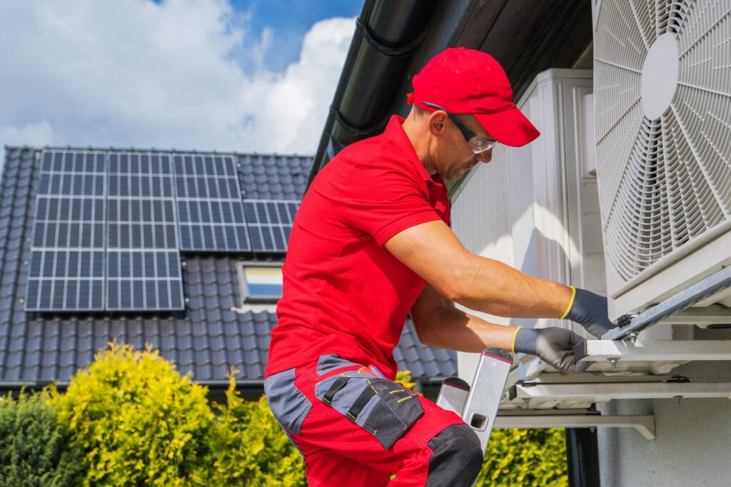 An HVAC professional dressed in red on a ladder installing a new residential HVAC unit.