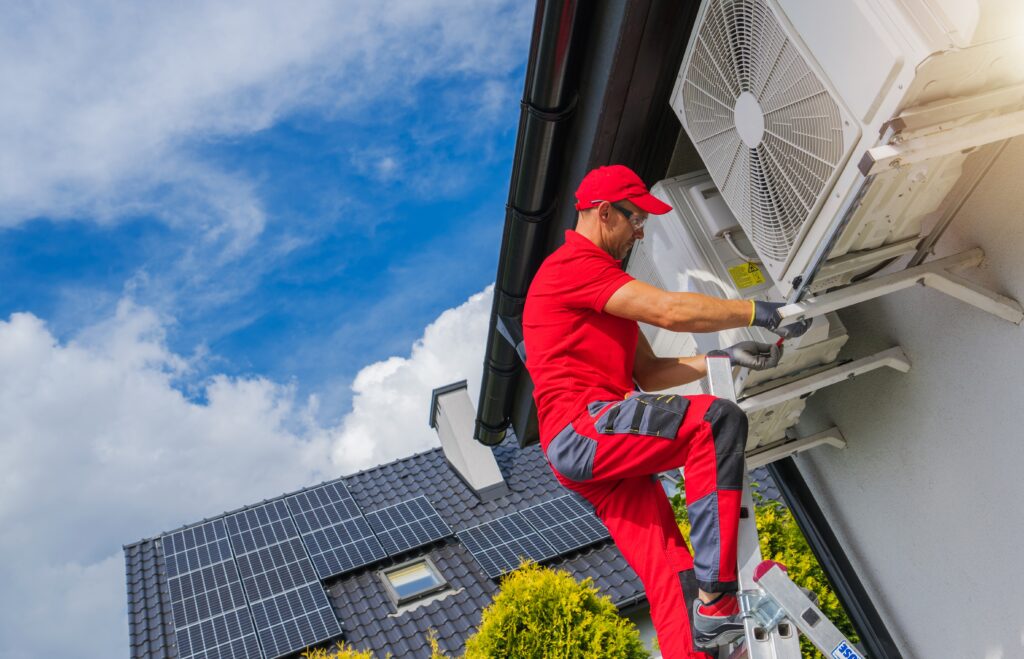 An HVAC technician dressed in red on top of a ladder repairing a residential HVAC unit.