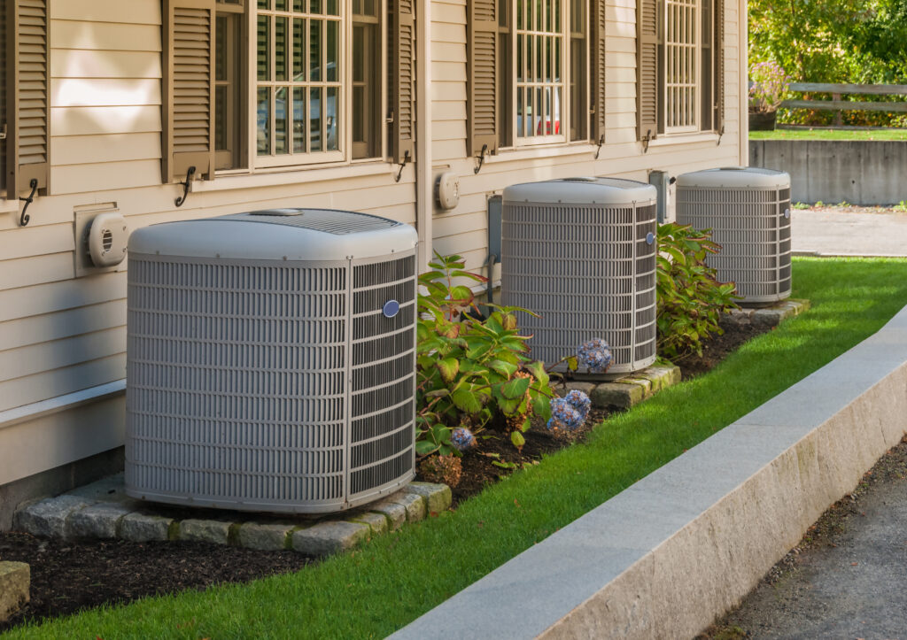An image of three residential HVAC units side-by-side on a grassy patch next to a home.