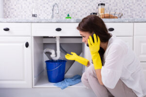 young woman calling plumber in front of sink with bucket catching leaking water
